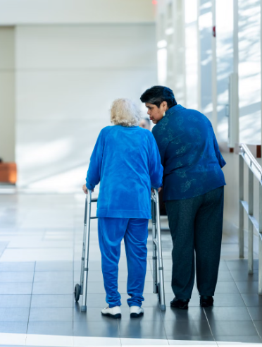 Caregiver walking closely beside an elderly woman using a walker in a bright indoor hallway