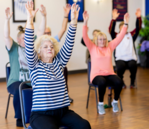 Group of elderly women doing a seated stretching exercise class with arms raised